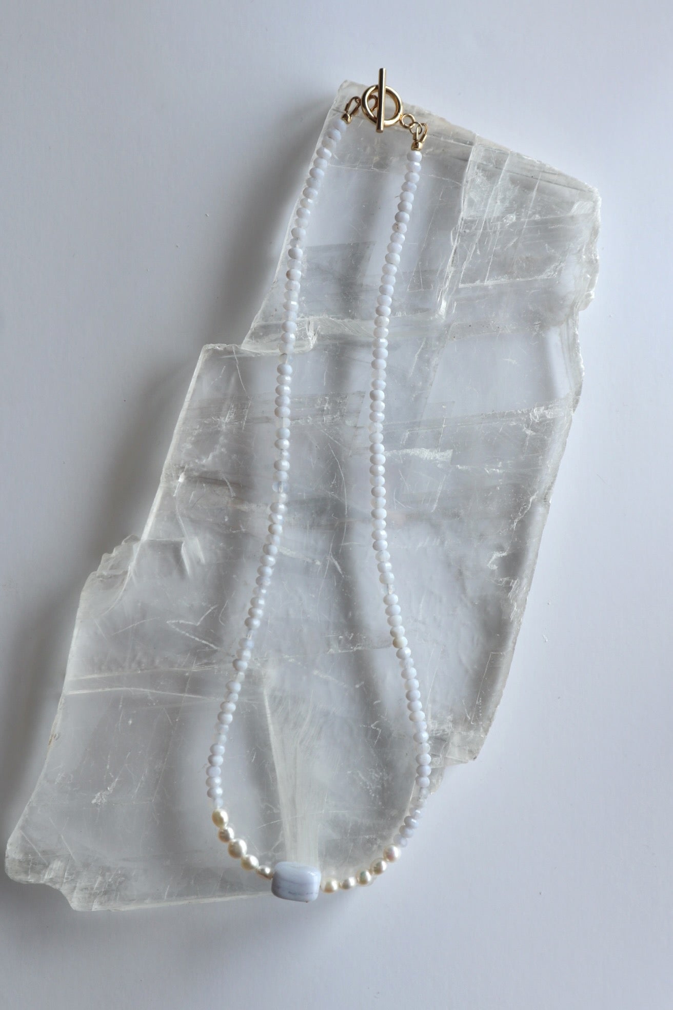Sky necklace by Auréad Designs laid flat on a selenite slab, showing the full length with blue lace agate rondelles, freshwater pearl accents, rectangular focal bead, and gold toggle clasp
