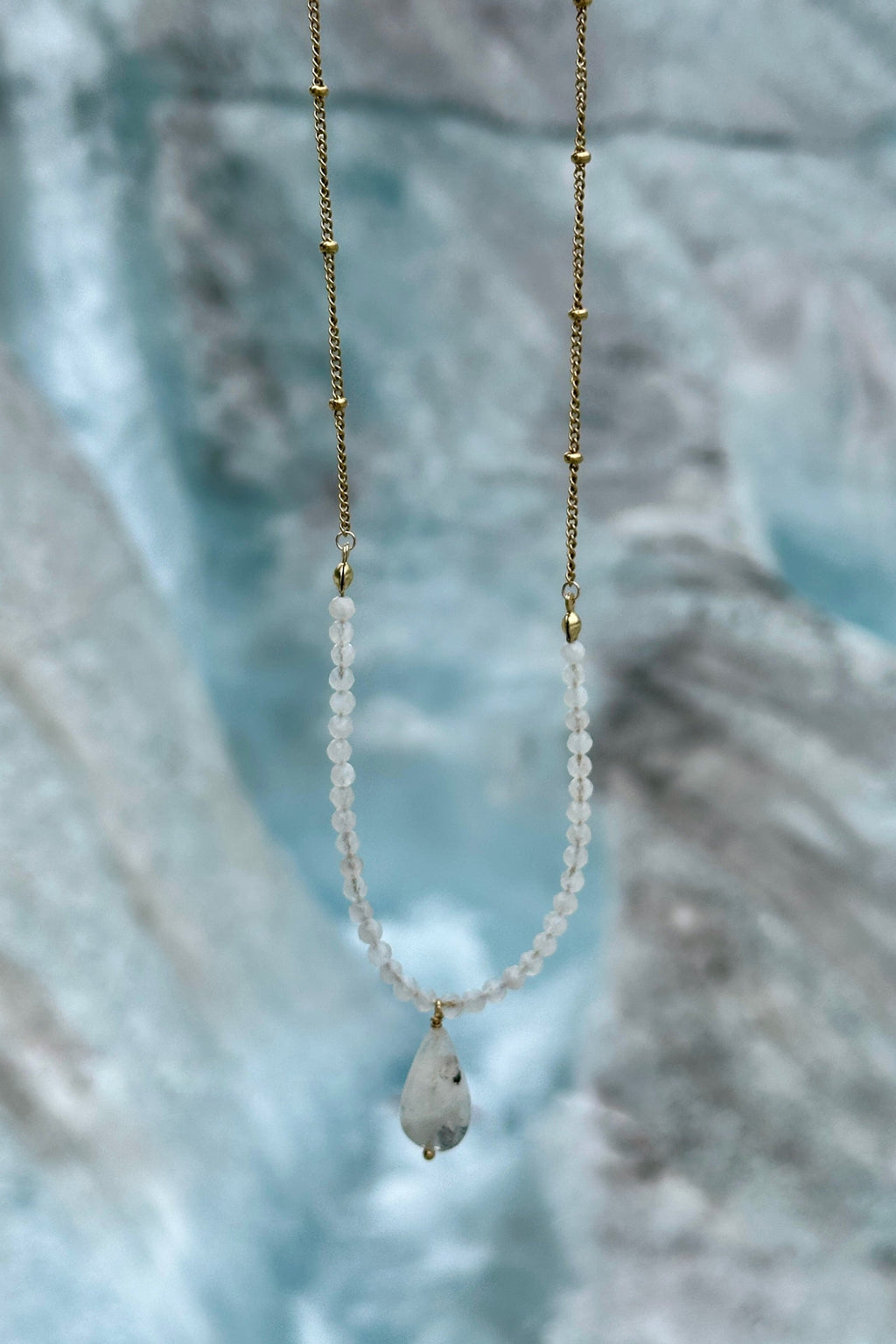 Snow necklace by Auréad Designs held against the blue ice face of the Athabasca Glacier, with rainbow moonstone rondelles, gold satellite chain, and moonstone teardrop pendant