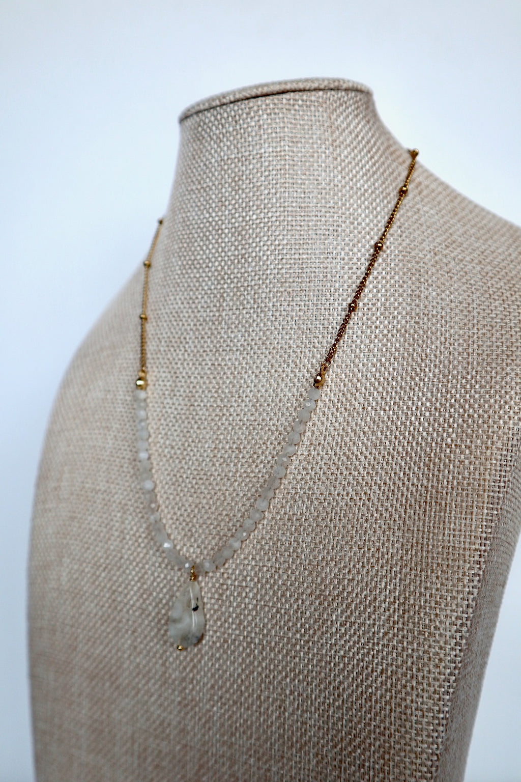 Snow necklace by Auréad Designs on a linen bust, angled view, showing the moonstone teardrop pendant and gold satellite chain