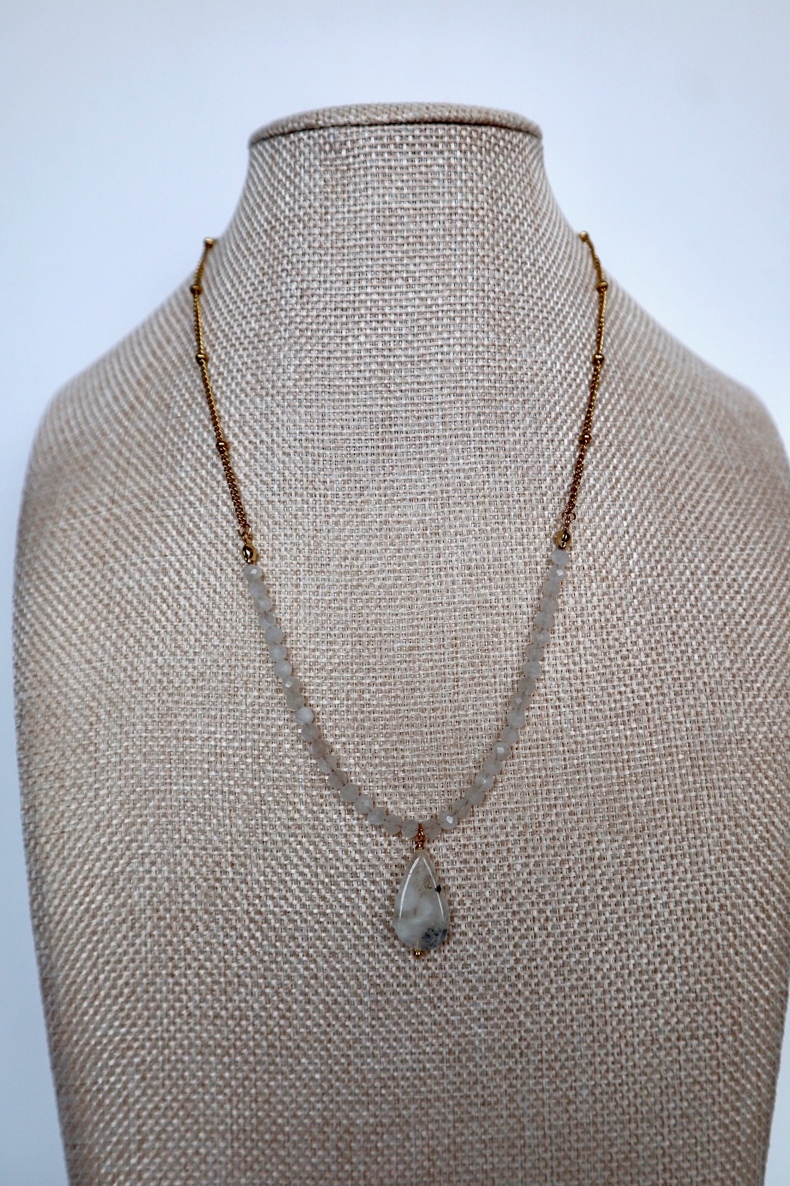 Snow necklace by Auréad Designs on a linen bust, front view, featuring rainbow moonstone rondelles on a gold satellite chain with a moonstone teardrop pendant
