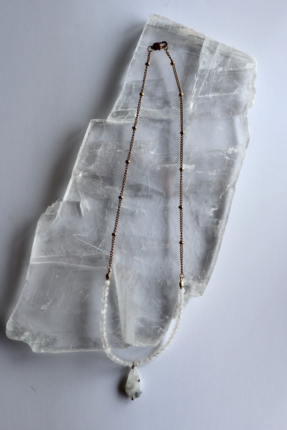 Snow necklace by Auréad Designs laid flat on a selenite slab, showing the full length with rainbow moonstone rondelles, gold satellite chain, moonstone teardrop pendant, and gold lobster clasp