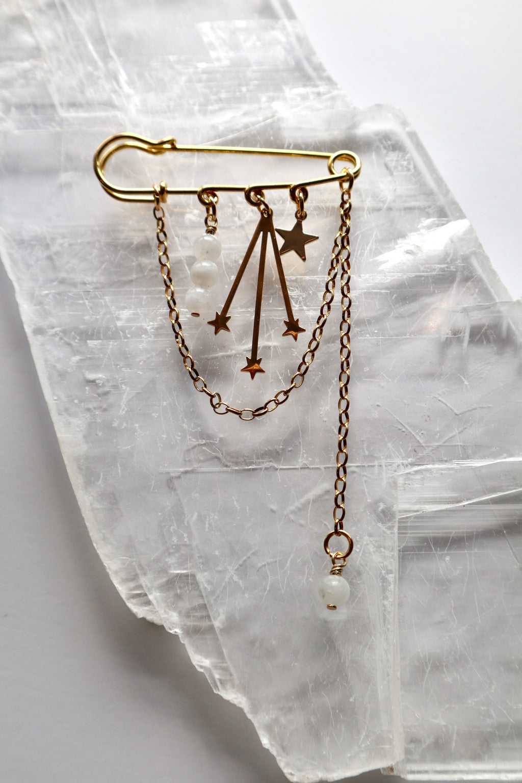 Stellune pin brooch by Auréad Designs, featuring moonstone beads, gold stainless steel charms, and draped gold chains, laid flat on a white selenite crystal