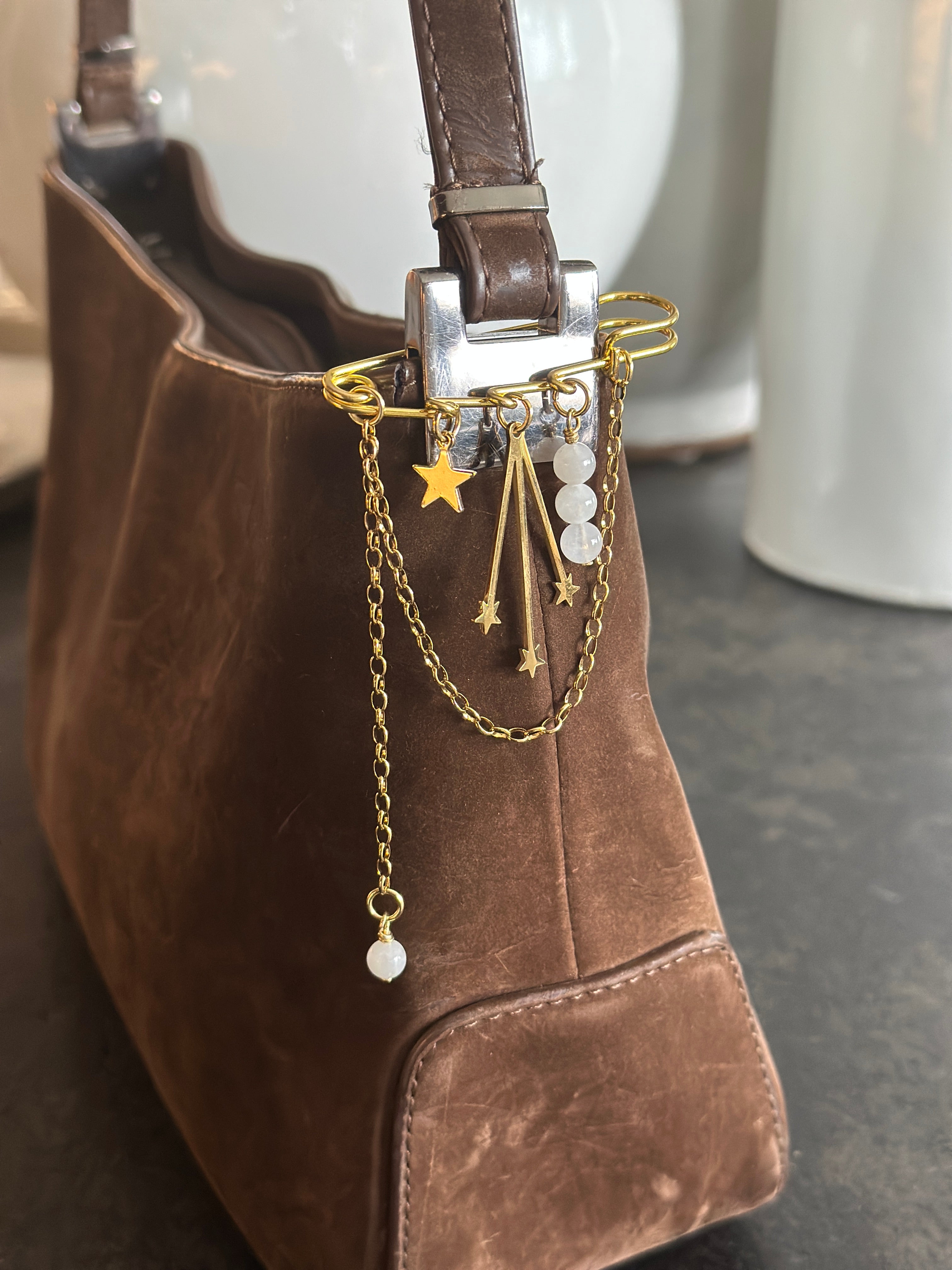 Stellune pin brooch by Auréad Designs pinned to a brown leather bag, with moonstone beads and gold chain drape visible against the warm-toned fabric