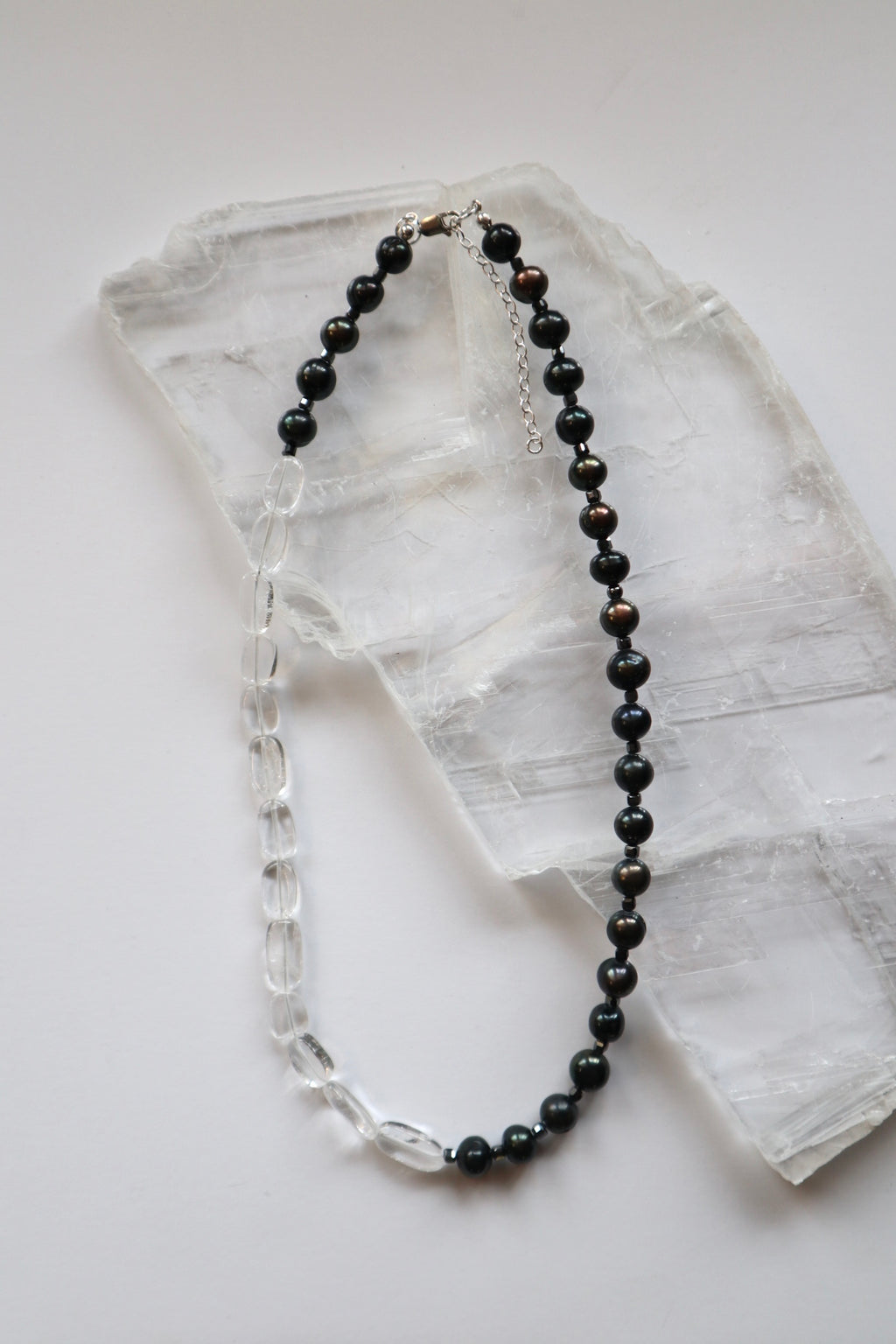 Tashira necklace by Auréad Designs, featuring clear quartz, black peacock freshwater pearls, and hematite spacer beads with a sterling silver clasp, laid flat on a selenite slab