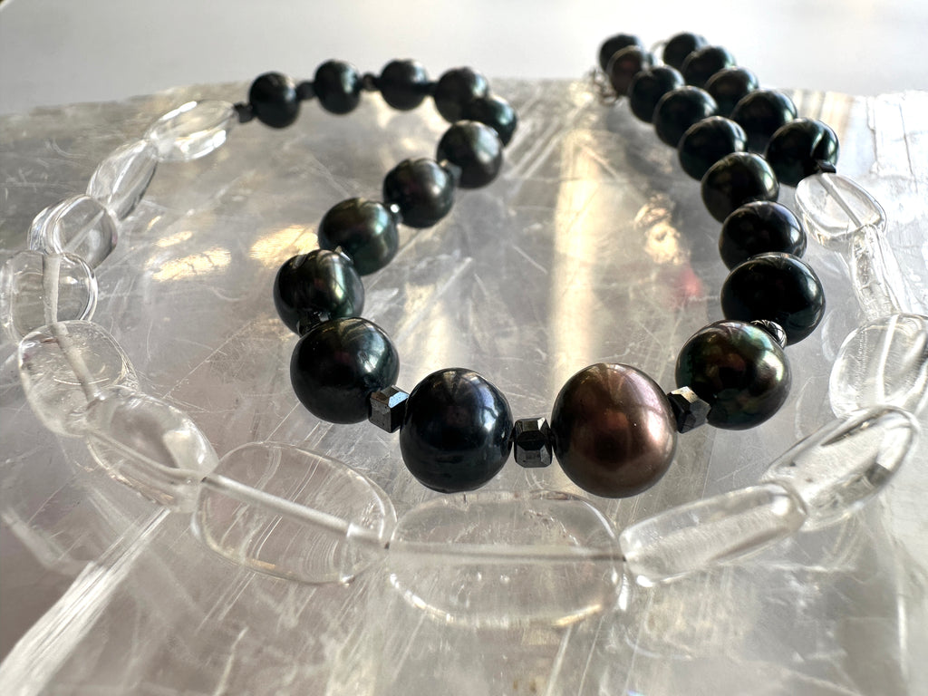 Tashira necklace by Auréad Designs displayed on a light surface, showcasing the full strand of clear quartz, black peacock freshwater pearls, and hematite spacer beads