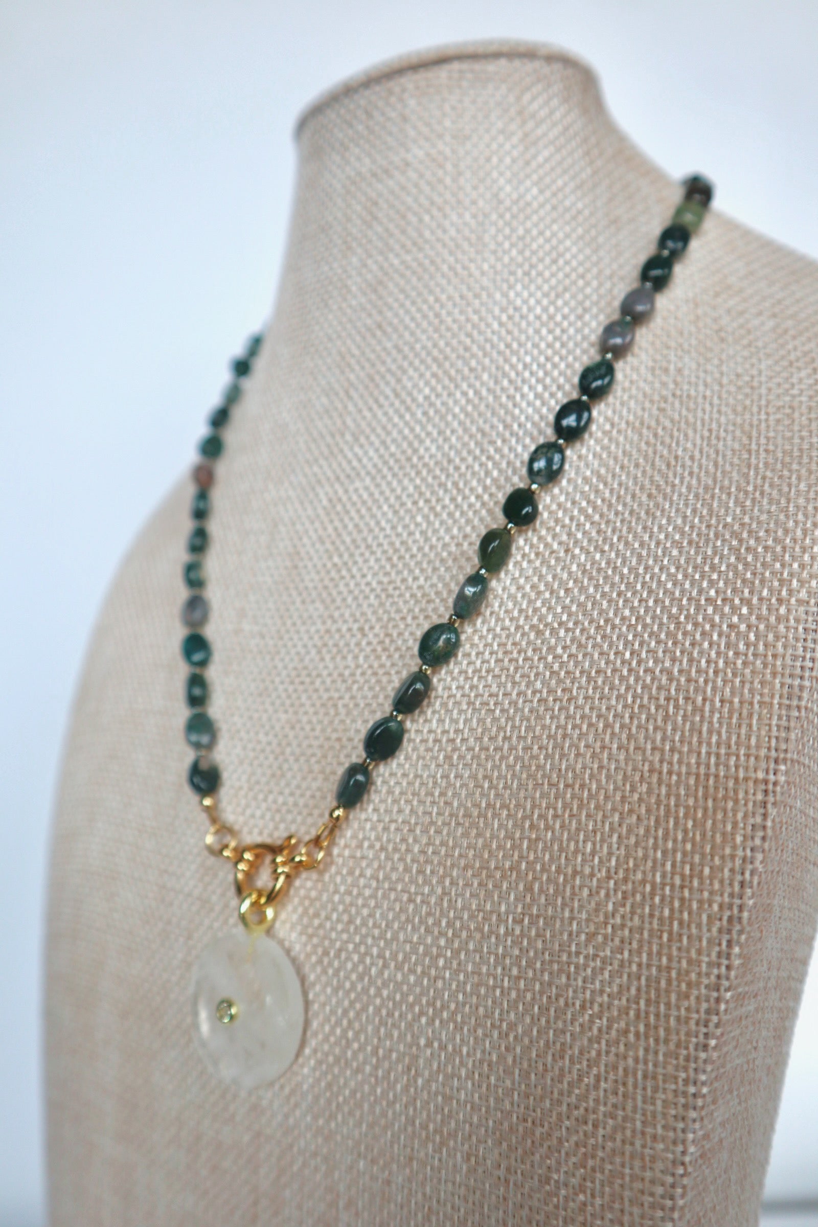 Trail necklace by Auréad Designs on a linen bust, angled view, showing the moss agate beads and clear quartz donut pendant