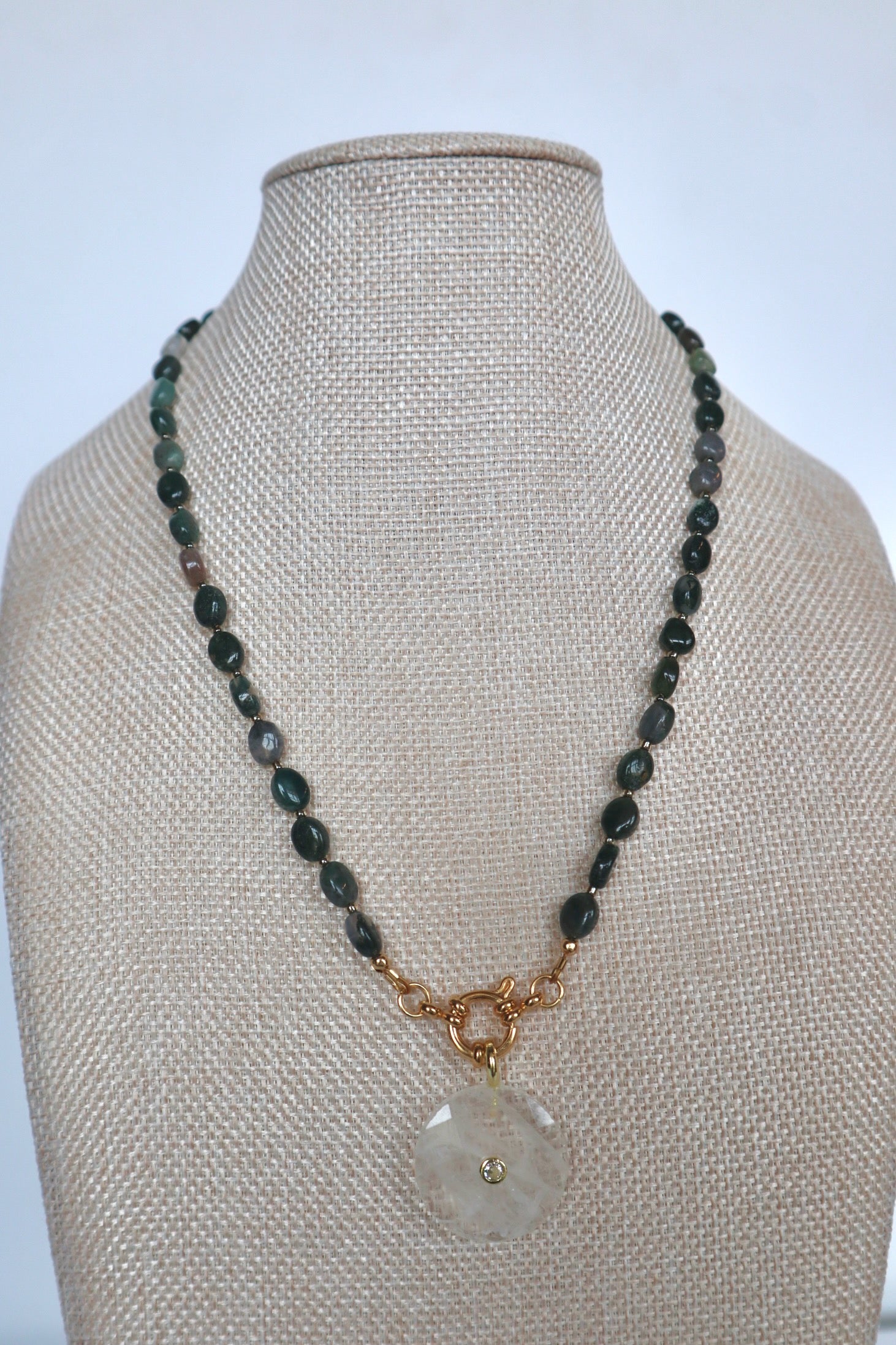 Trail necklace by Auréad Designs on a linen bust, front view, featuring moss agate oval beads and a clear quartz donut pendant with a gold ring clasp