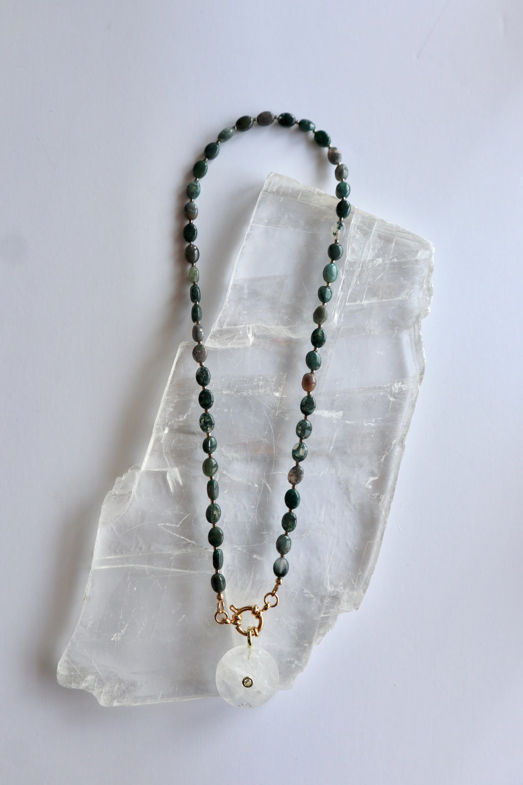 Trail necklace by Auréad Designs laid flat on a selenite slab, showing the full length with moss agate oval beads, gold ring clasp, and clear quartz donut pendant