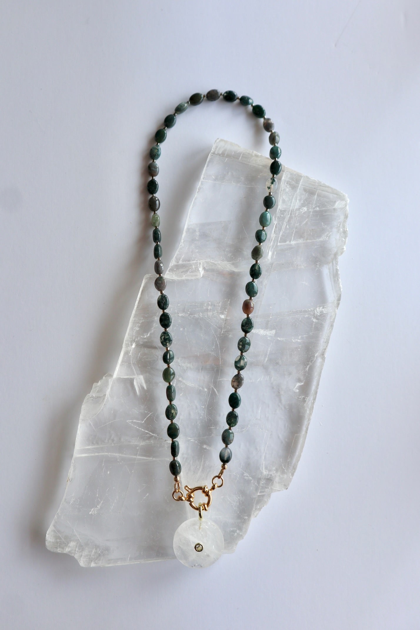 Trail necklace by Auréad Designs laid flat on a selenite slab, showing the full length with moss agate oval beads, gold ring clasp, and clear quartz donut pendant