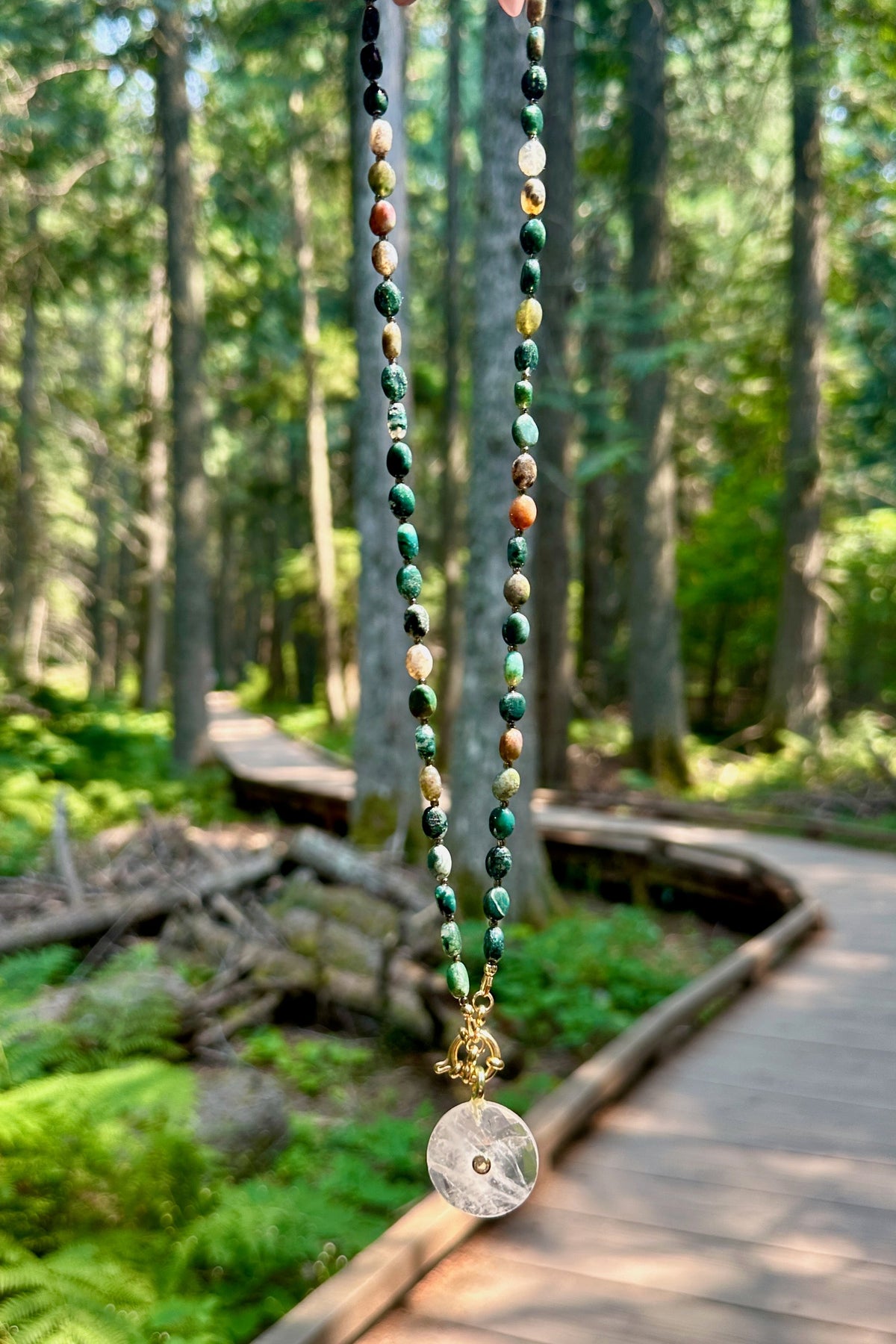 Trail necklace by Auréad Designs held against the cedar forest boardwalk of the Trail of the Cedars in Glacier National Park, with moss agate oval beads and a clear quartz donut pendant