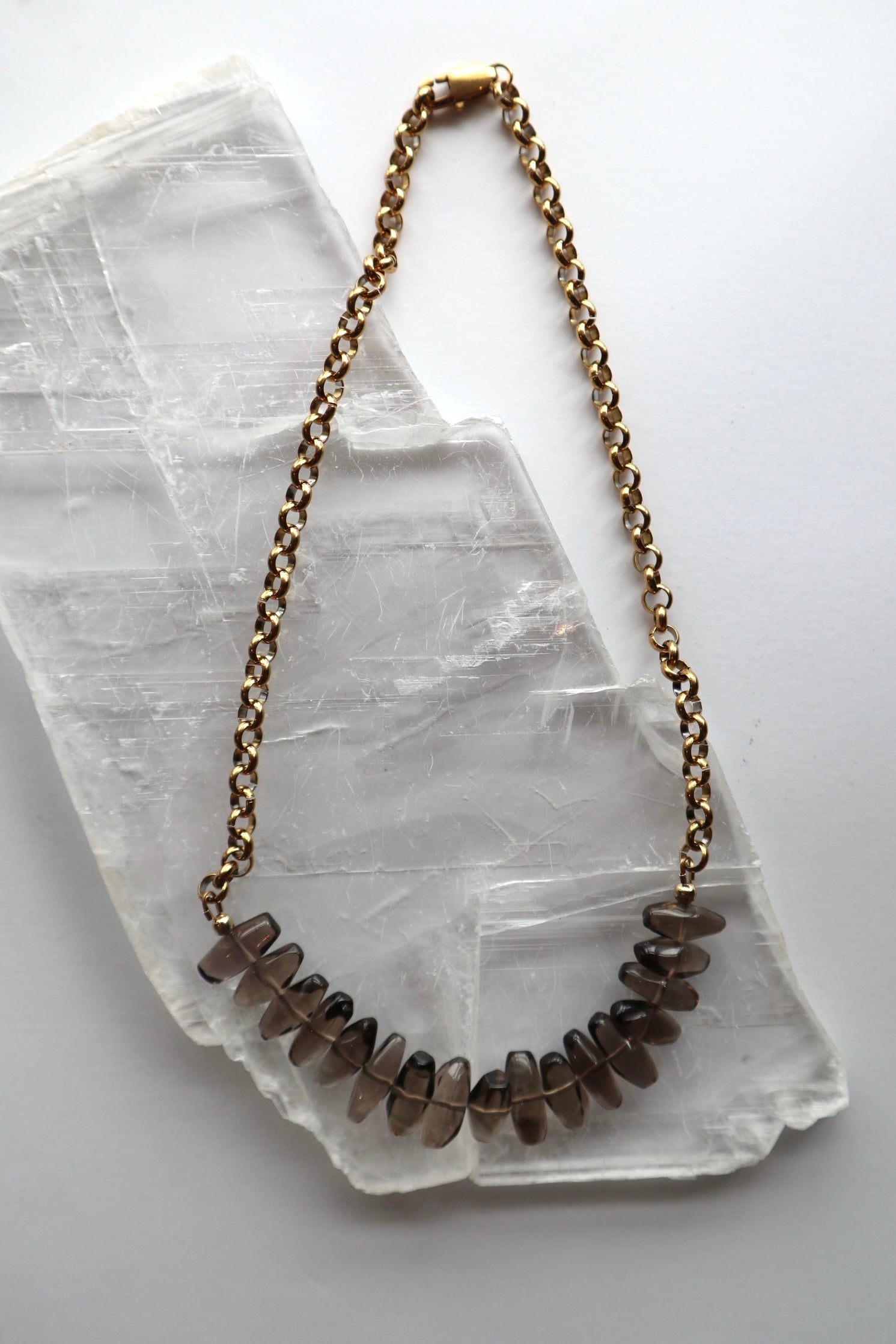 Umbra necklace by Auréad Designs, featuring faceted smoky quartz drops on a gold stainless steel rolo chain, laid flat on a selenite slab