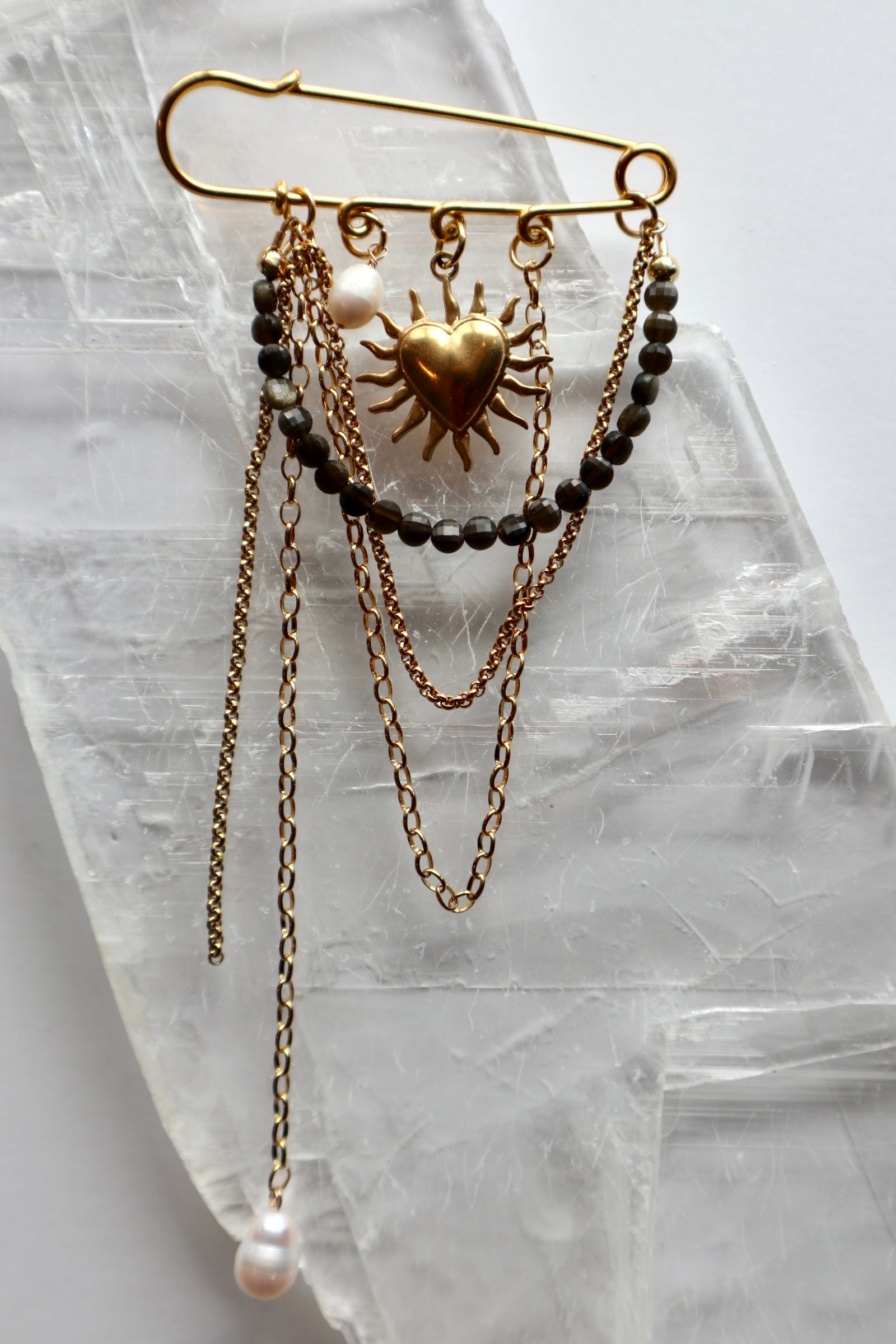 Velora pin brooch by Auréad Designs, featuring gold sheen obsidian, freshwater pearl, and gold stainless steel chains with a heart charm, laid flat on a white selenite crystal