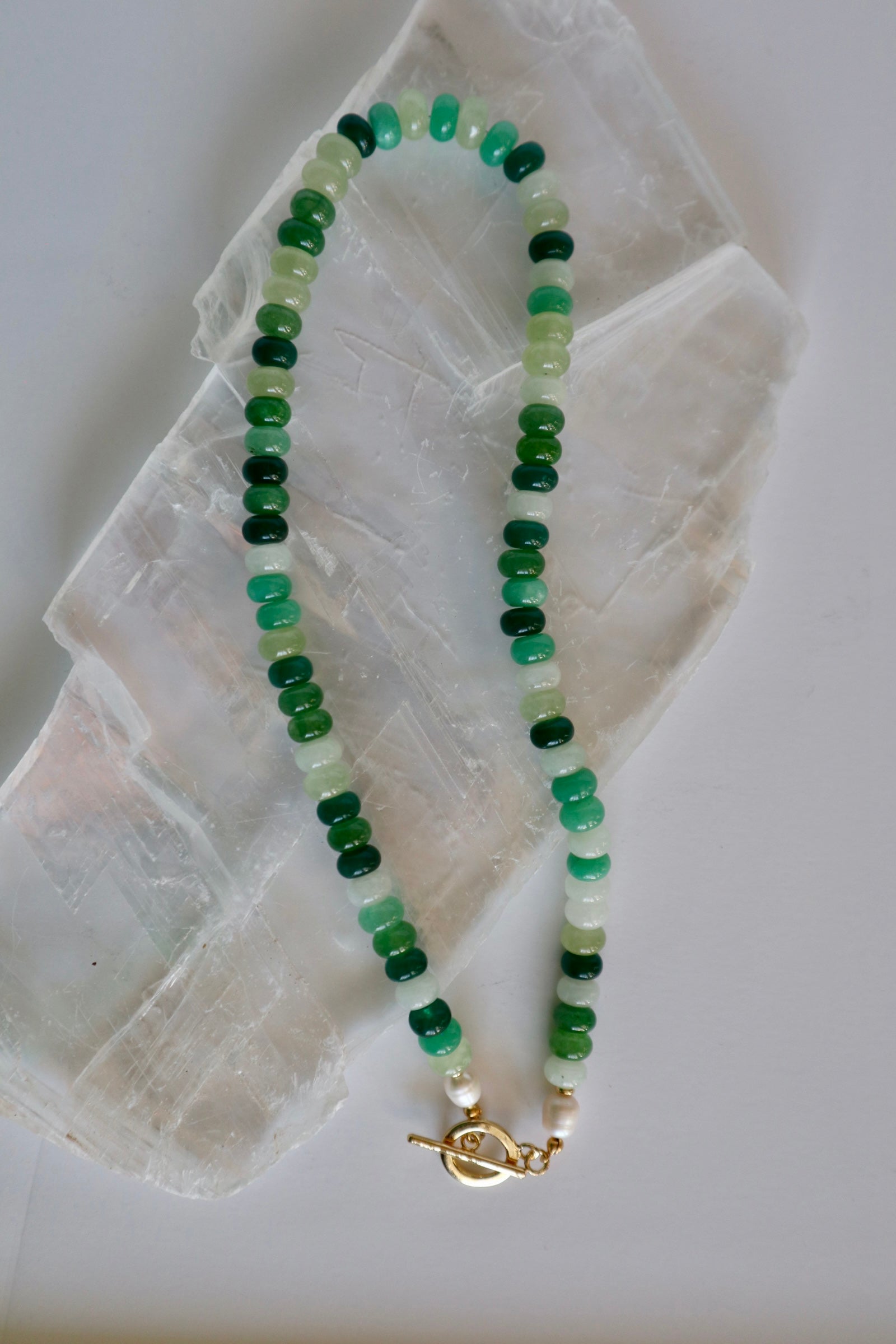 Verdelle necklace by Auréad Designs, featuring jade and freshwater pearl beads with a gold toggle clasp, laid flat on a white selenite crystal