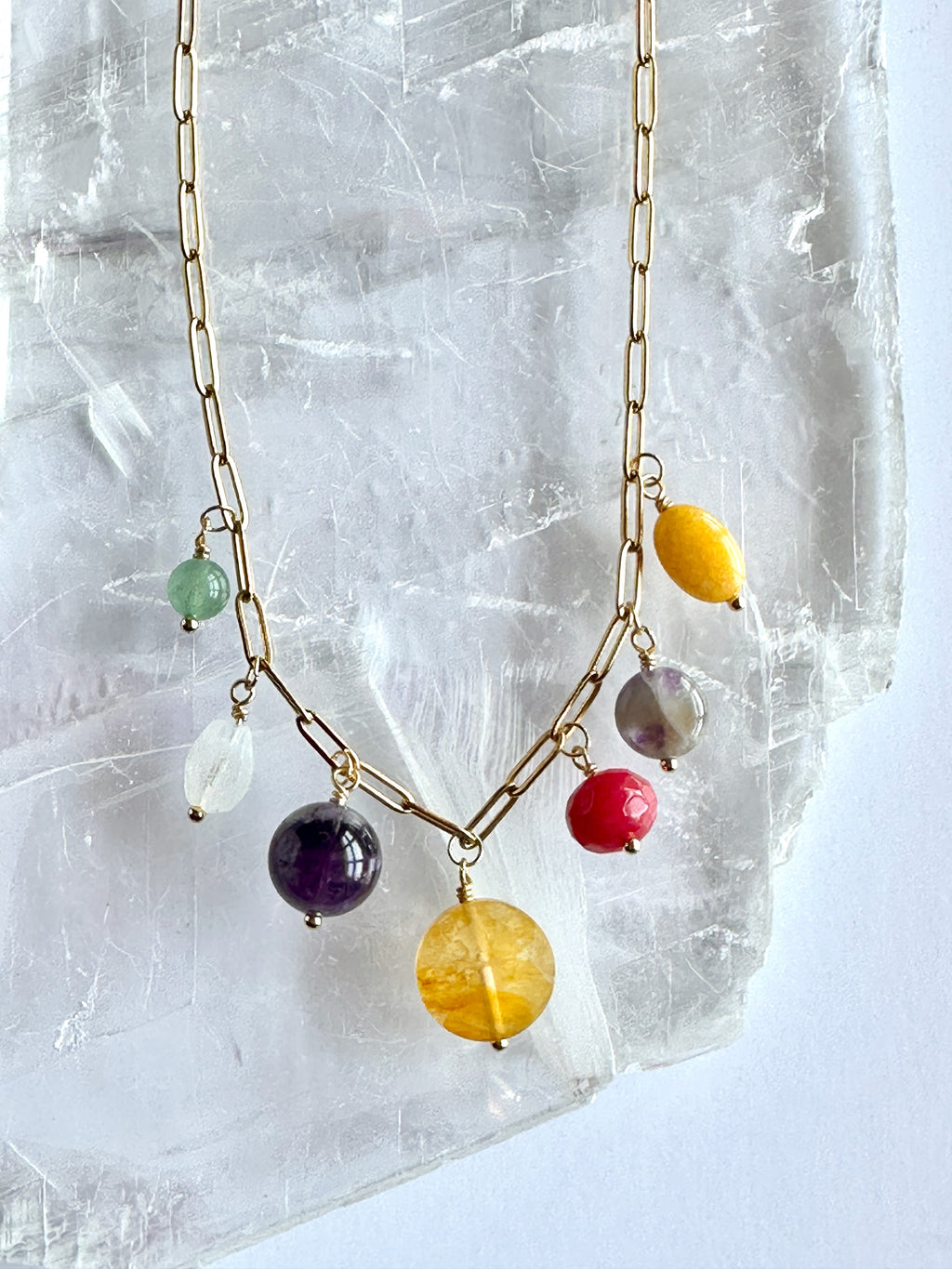 Close-up of the colorful gemstone drop cluster on the Wildflower necklace by Auréad Designs against a selenite slab, showing green aventurine, amethyst, yellow jade, golden healer quartz, rainbow moonstone, fuschia-dyed agate, and freshwater pearl