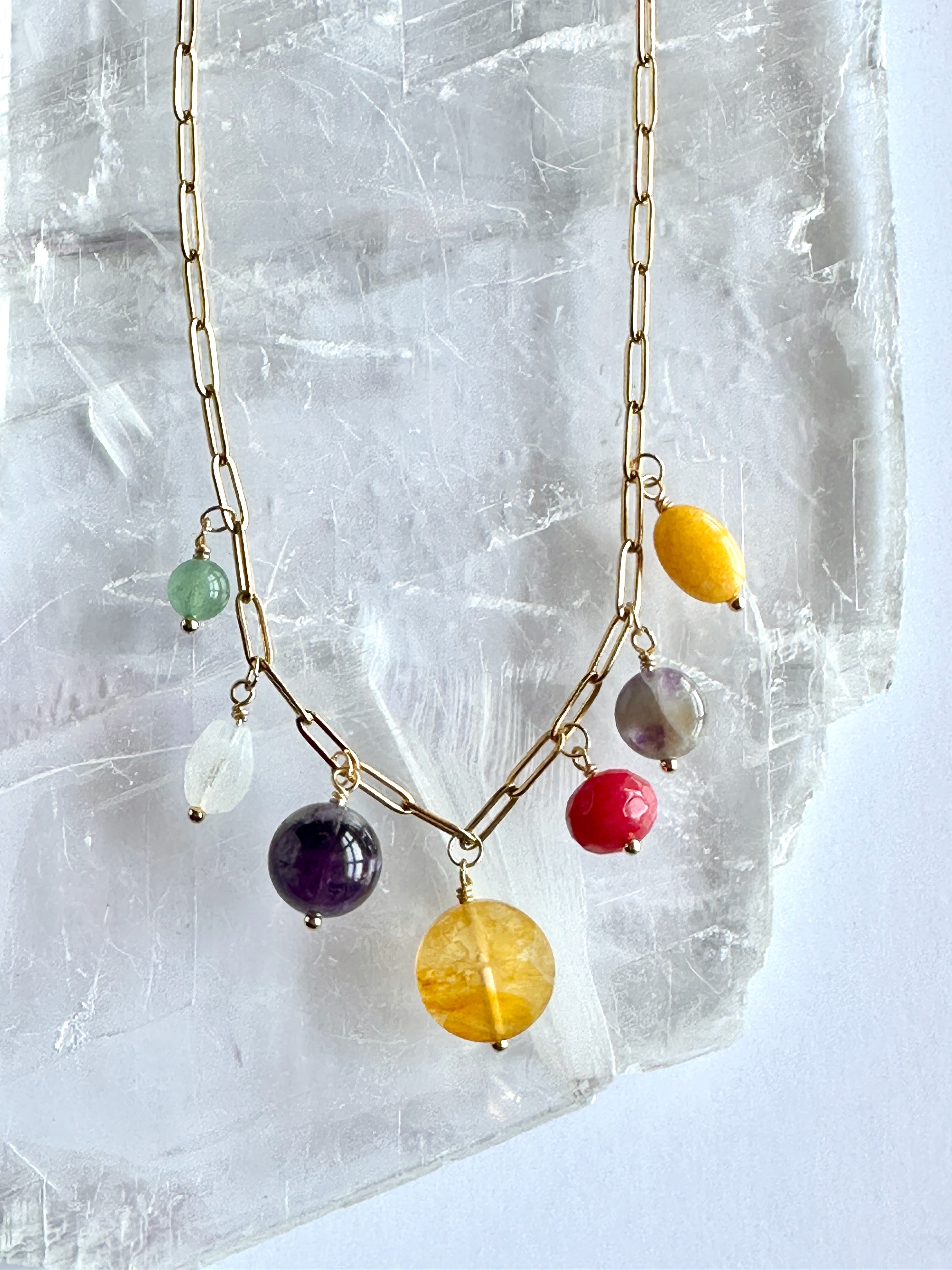 Close-up of the colorful gemstone drop cluster on the Wildflower necklace by Auréad Designs against a selenite slab, showing green aventurine, amethyst, yellow jade, golden healer quartz, rainbow moonstone, fuschia-dyed agate, and freshwater pearl