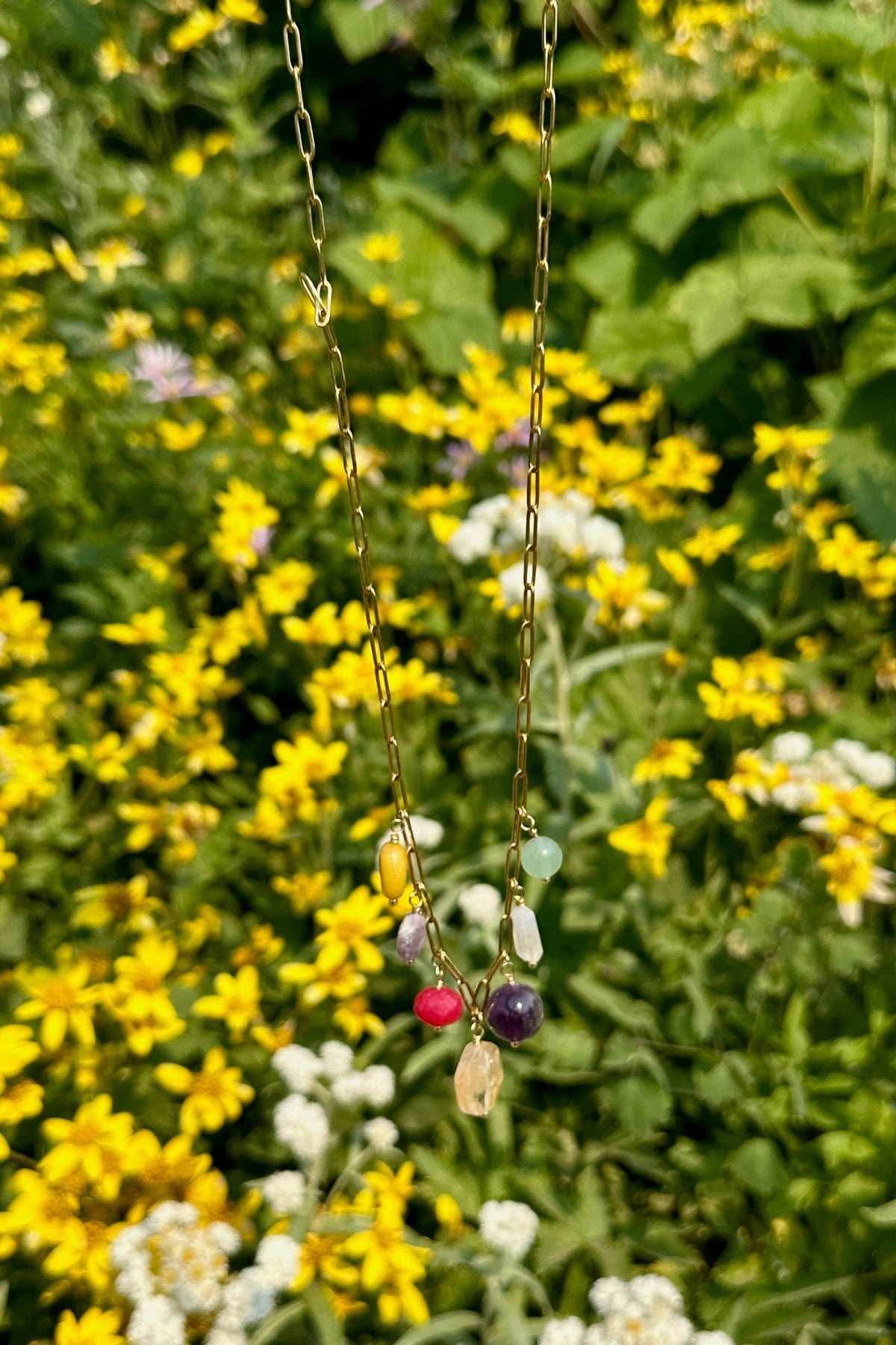 Wildflower necklace by Auréad Designs hanging in a field of yellow wildflowers along Going-to-the-Sun Road in Glacier National Park, showing the cluster of colorful gemstone drops on a gold paperclip chain