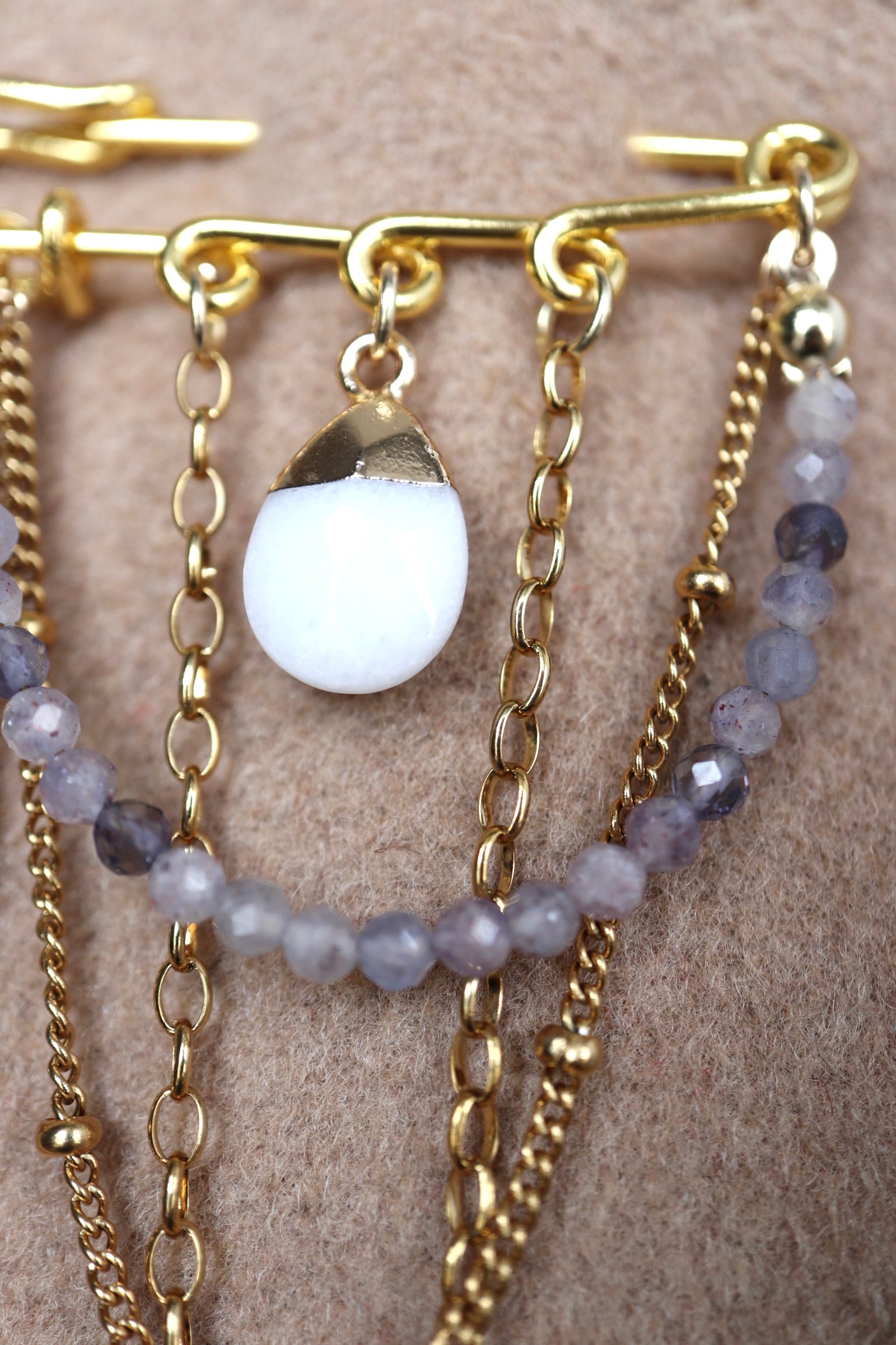 Close-up of Celune's gold safety pin bar on camel wool showing the pin mechanism, jump rings, and how the chains and iolite strand attach to the bar structure