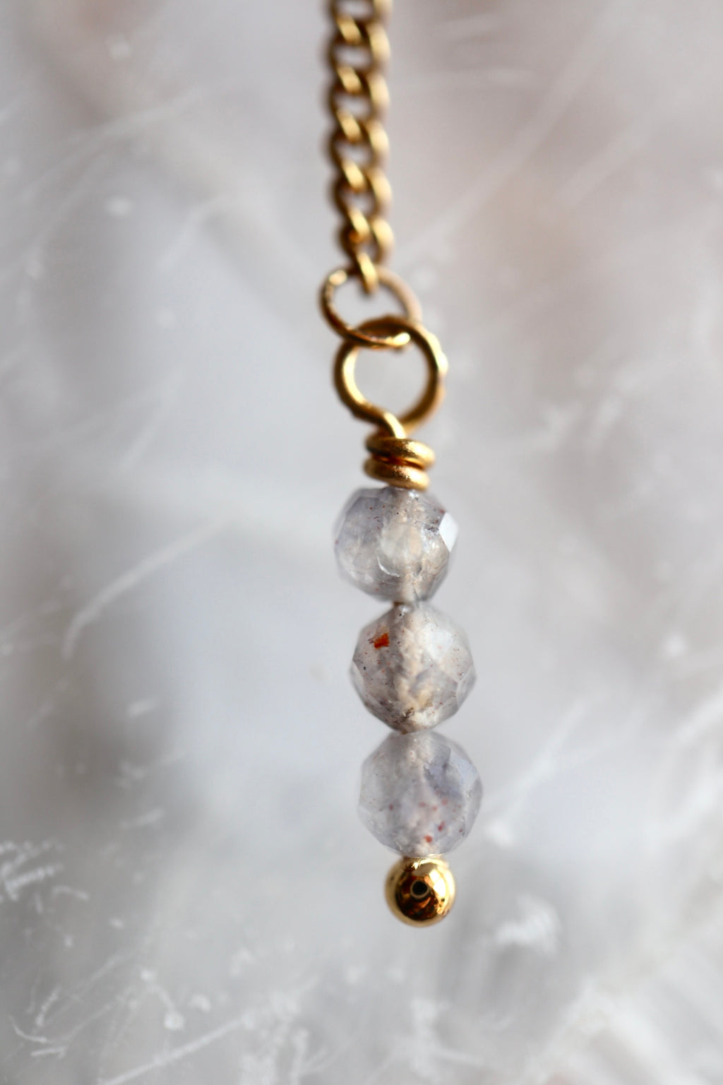 Close-up of Celune's iolite bead tassel drop showing three stacked faceted iolite beads wire-wrapped onto a gold chain with a gold bead end cap, against a selenite background