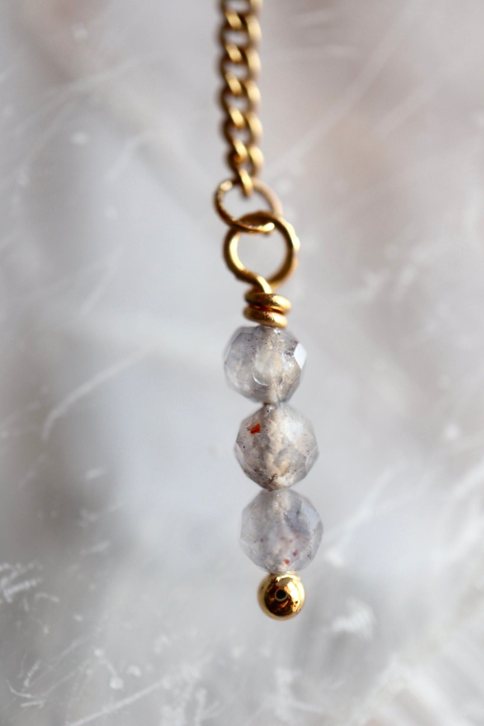 Close-up of Celune's iolite bead tassel drop showing three stacked faceted iolite beads wire-wrapped onto a gold chain with a gold bead end cap, against a selenite background