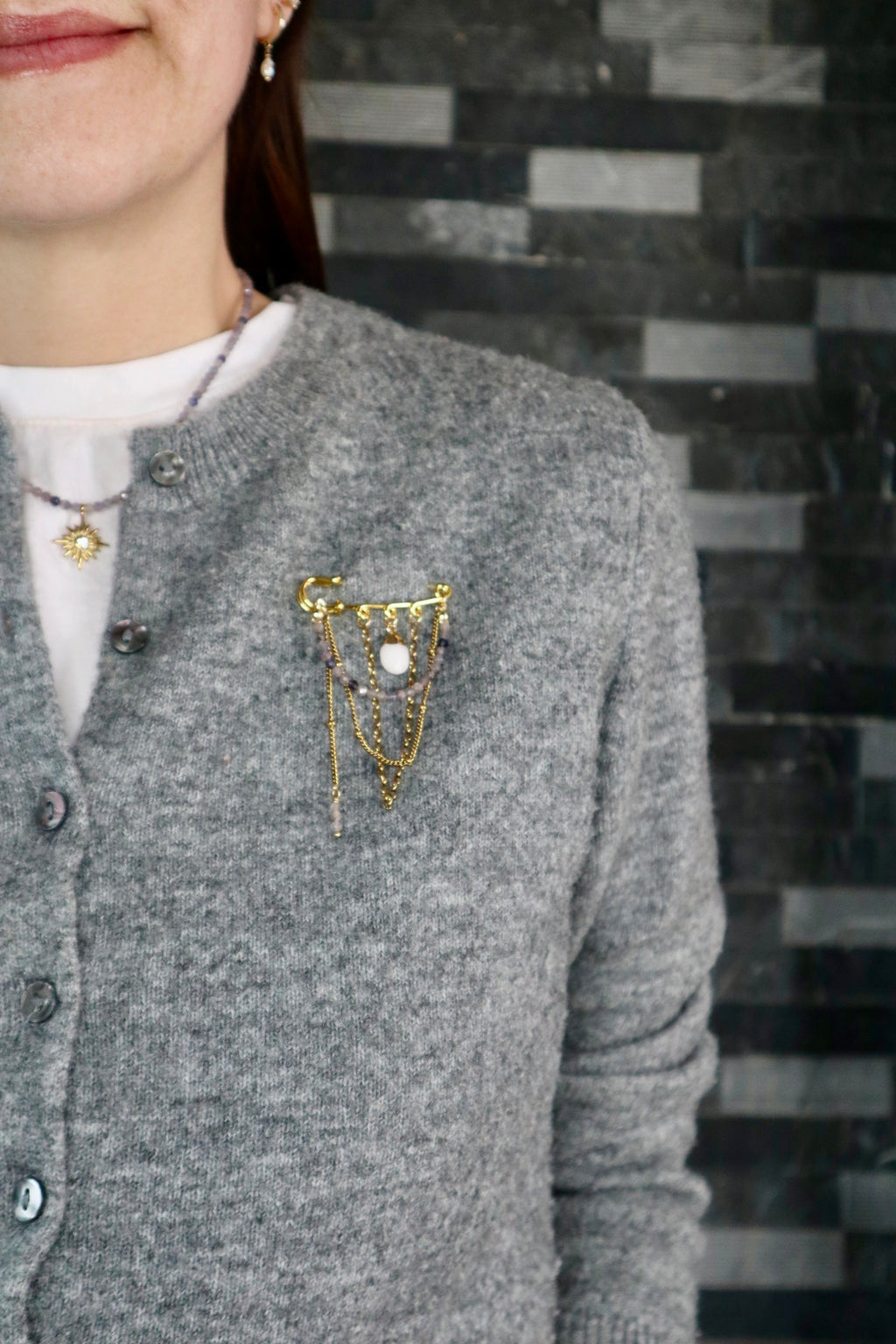 Celune iolite chain pin brooch pinned to the chest of a grey knit cardigan, showing the draped gold chain and white jade charm movement in a styled outfit