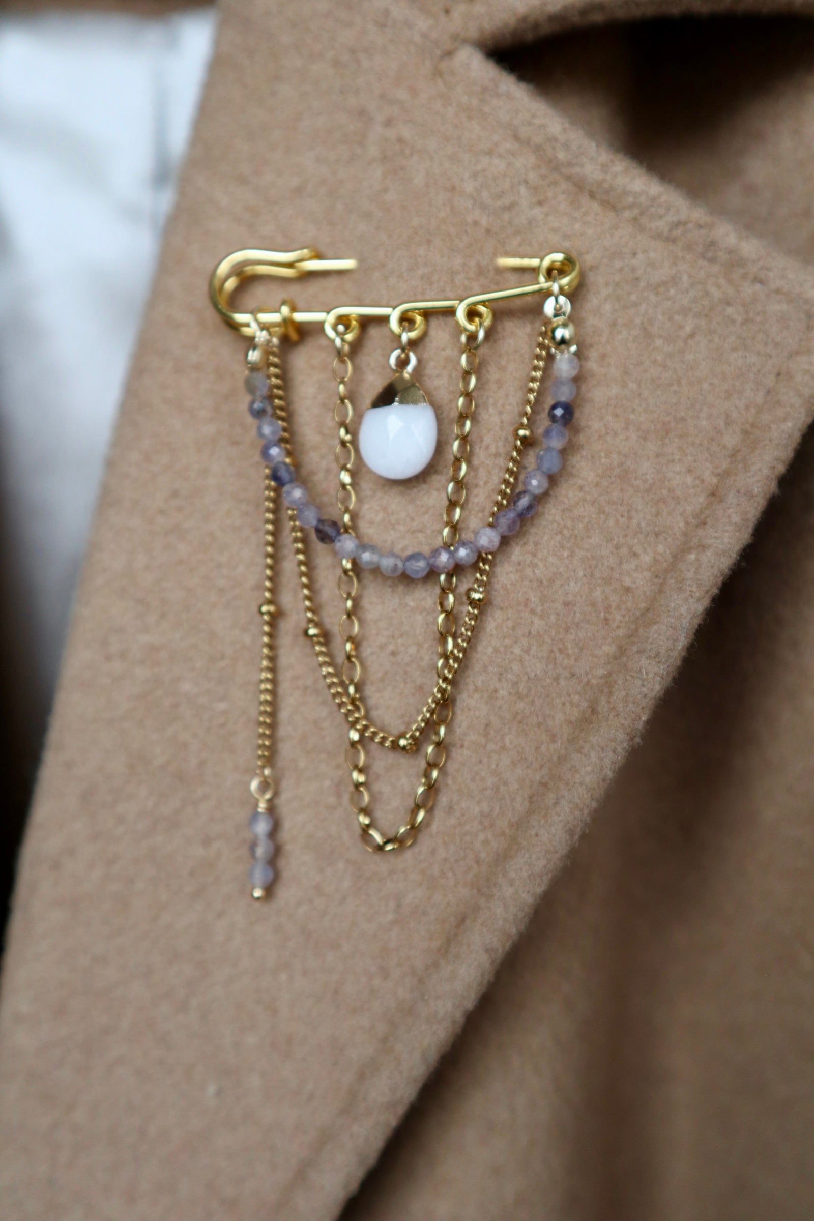 Celune brooch pinned to the lapel of a camel wool coat, showing the full layered gold chain drape, iolite bead strand, and white jade charm against a warm tan fabric