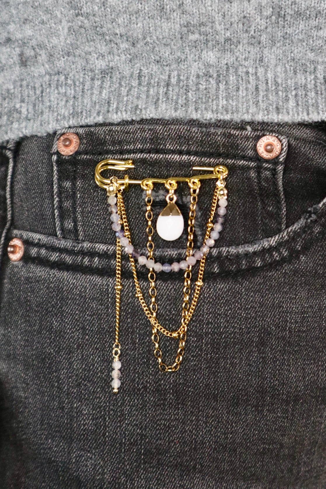Celune iolite chain pin brooch clipped to the pocket of dark wash jeans with a grey knit top, showing a casual everyday styling option with the full chain drape visible
