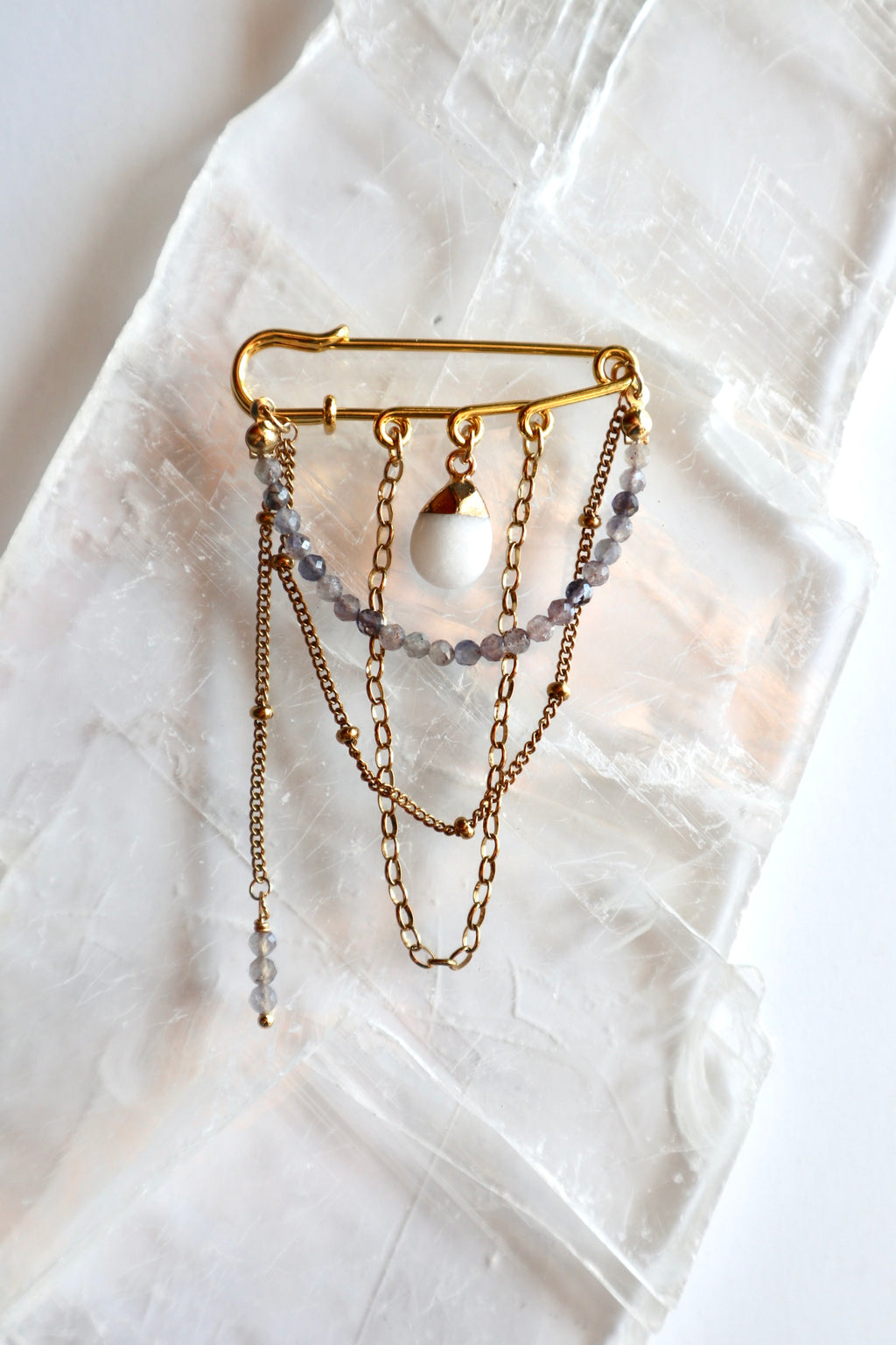Celune iolite and white jade chain pin brooch laid flat on a raw selenite slab, showing full design with draped gold chains and safety pin structure