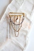 Celune iolite and white jade chain pin brooch laid flat on a raw selenite slab, showing full design with draped gold chains and safety pin structure