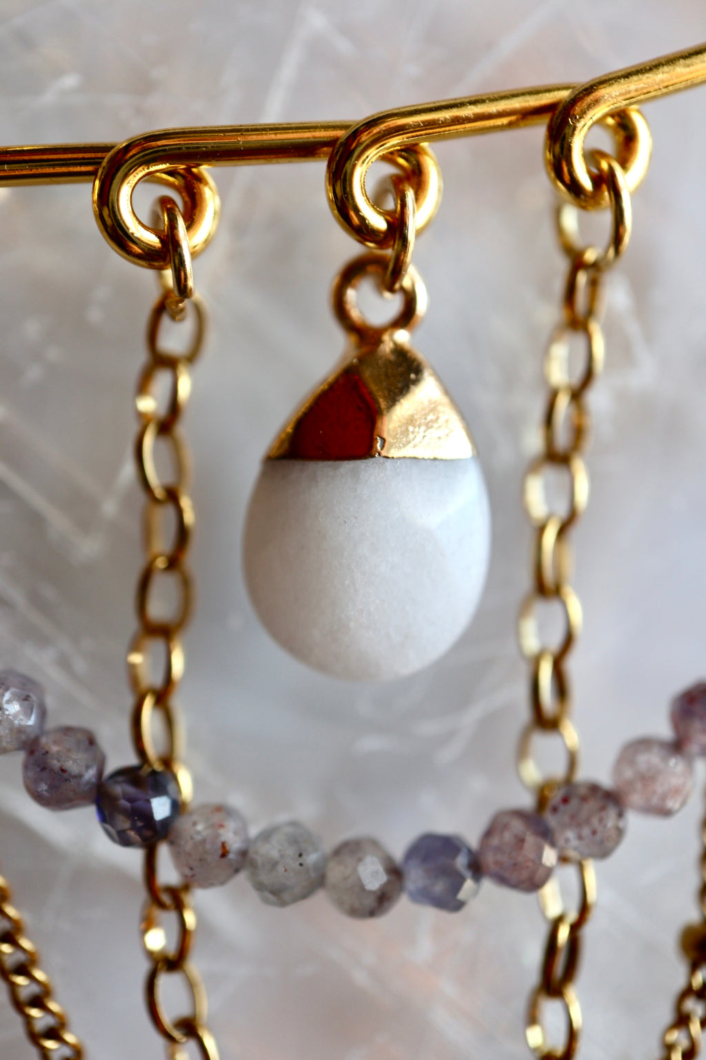 Macro close-up of Celune's white jade teardrop charm with gold cap, suspended from the safety pin bar alongside iolite beads and layered gold chains on a linen surface