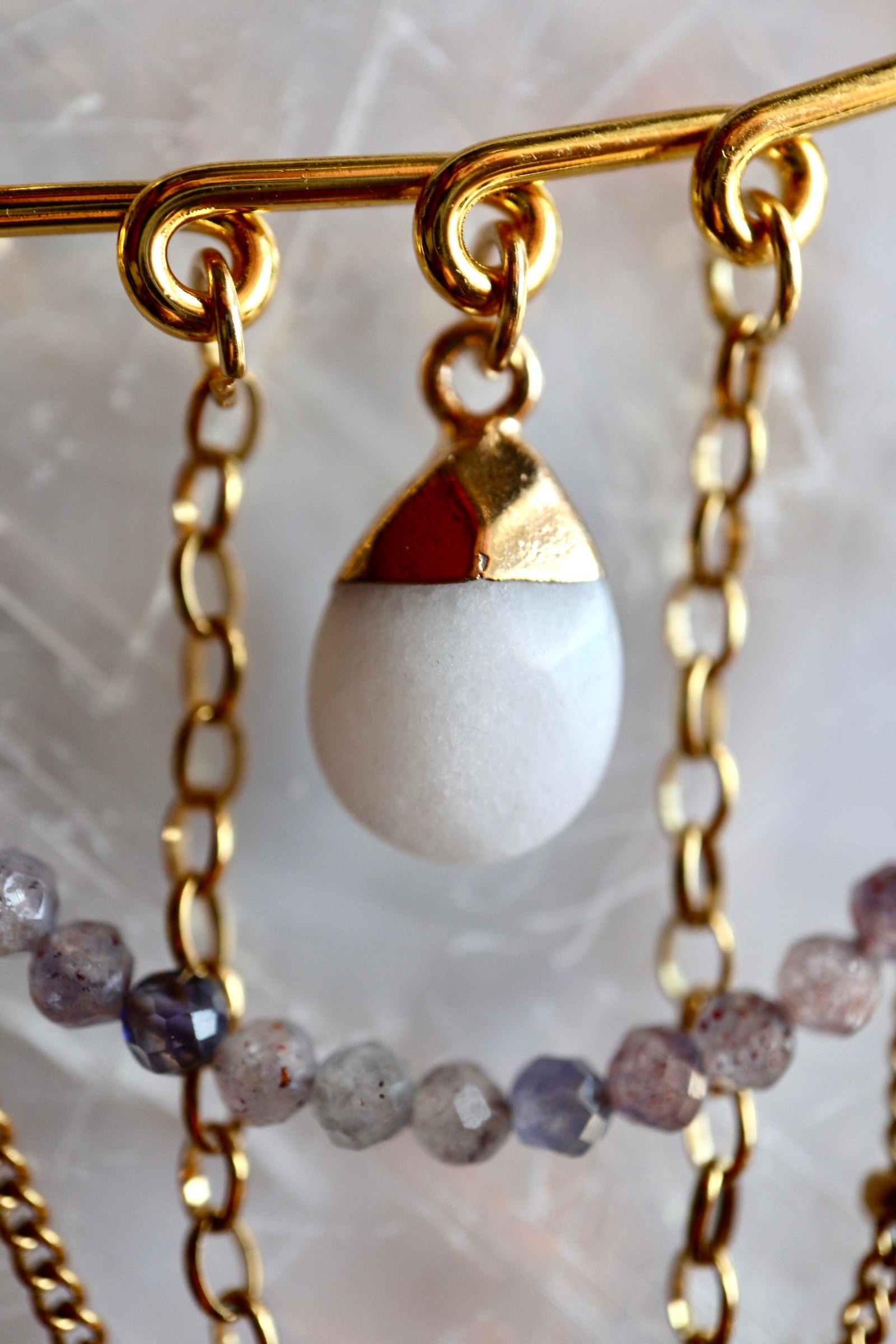 Macro close-up of Celune's white jade teardrop charm with gold cap, suspended from the safety pin bar alongside iolite beads and layered gold chains on a linen surface