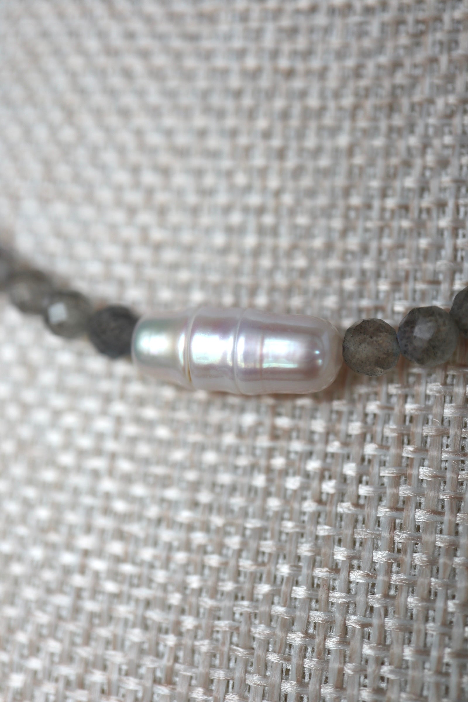 Macro close-up of Liora's centered freshwater pearl showing its lustrous iridescent surface flanked by faceted labradorite beads on a linen bust