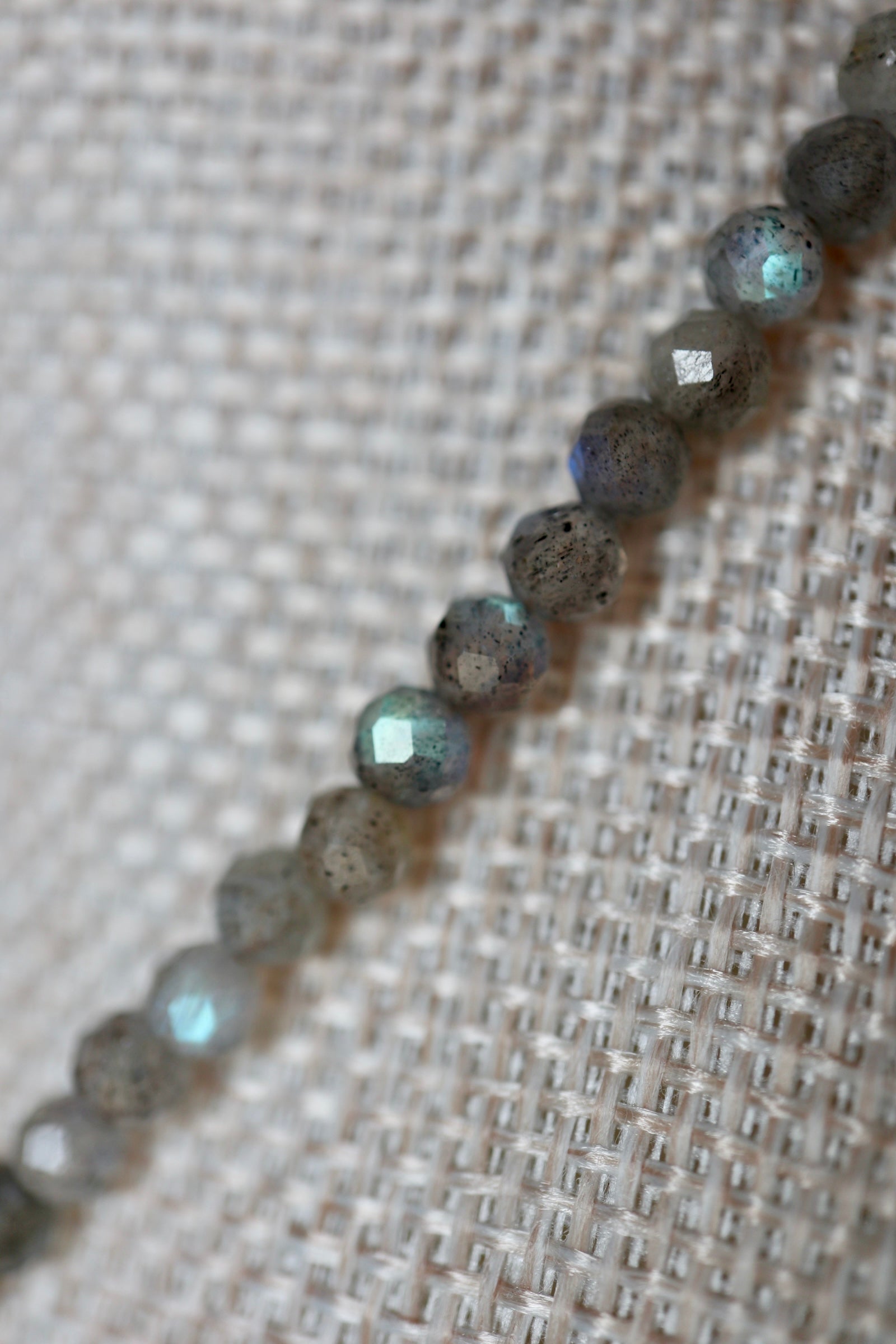 Extreme close-up of Liora's faceted labradorite beads on a linen bust, showing the characteristic blue and teal flash of labradorescence across the stone surfaces