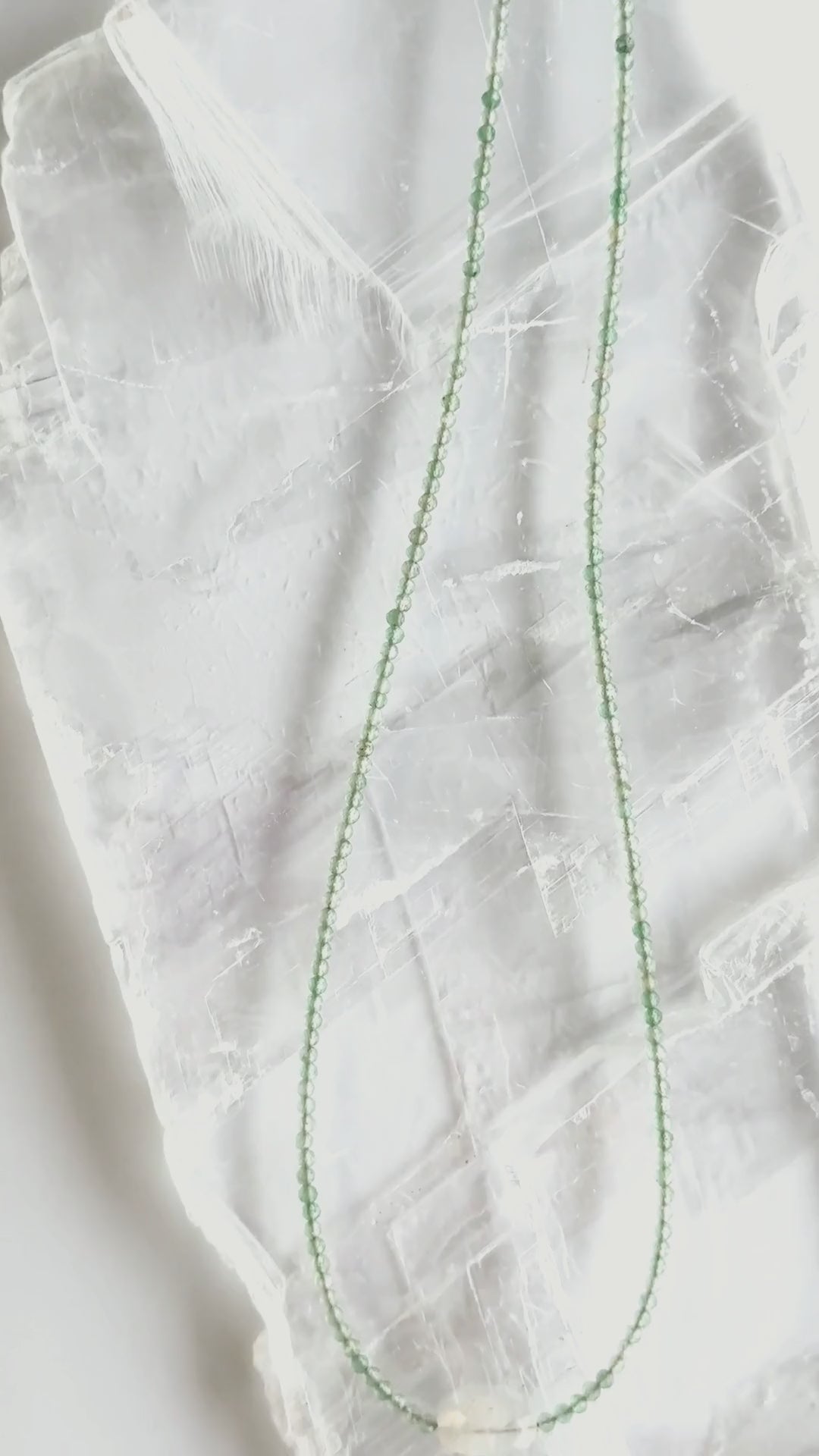 Video showcasing the Auréad Designs Primelle necklace featuring 2mm faceted green aventurine beads and a moonstone focal bead, shown on a linen bust form, worn on the neck layered over a white ribbed top, and styled on a white selenite slab with close-up detail of the beads and focal stone