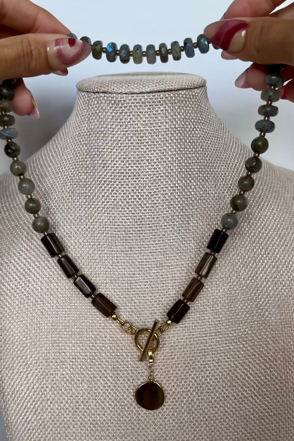 Video of the Ridge necklace by Auréad Designs, featuring labradorite rondelles and smoky quartz tube beads with a gold compass charm toggle clasp, shown in full, styled on a linen bust, and worn