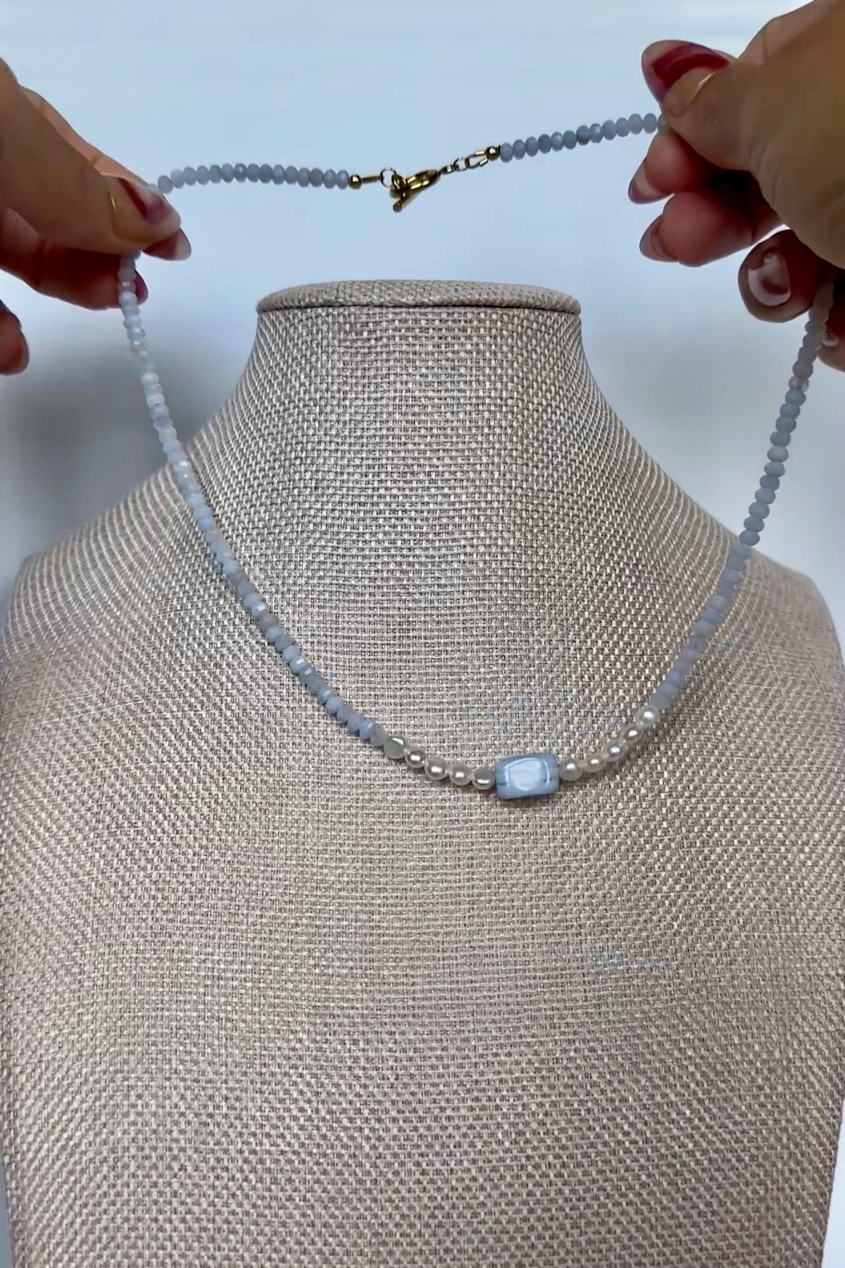 Video of the Sky necklace by Auréad Designs, featuring blue lace agate rondelles and freshwater pearl accents with a rectangular blue lace agate focal bead and gold toggle clasp, shown in full, styled on a linen bust, and worn