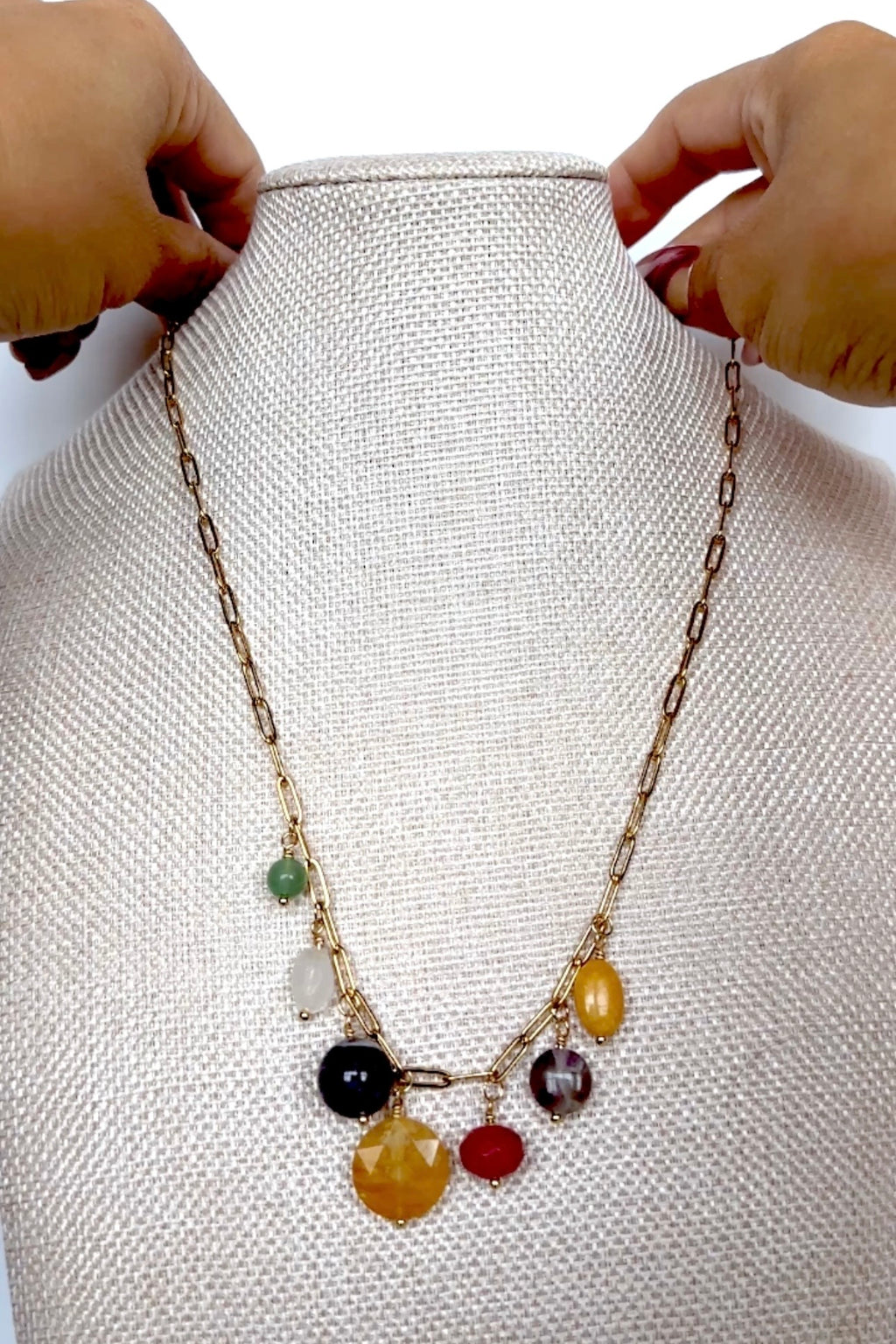 Video of the Wildflower necklace by Auréad Designs, featuring seven colorful gemstone drops including green aventurine, amethyst, yellow jade, golden healer quartz, rainbow moonstone, fuchsia-dyed agate, and freshwater pearl on a gold paperclip chain, shown in full, styled on a linen bust, and worn