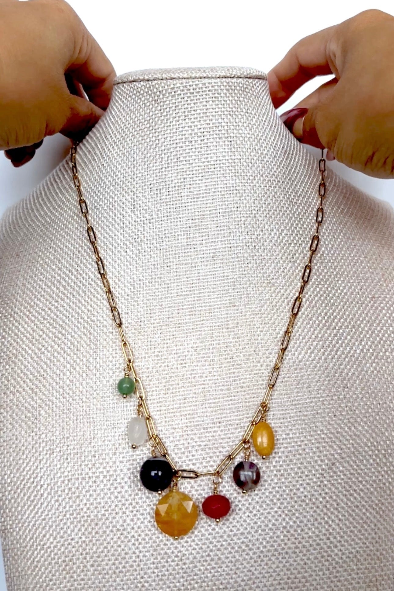 Video of the Wildflower necklace by Auréad Designs, featuring seven colorful gemstone drops including green aventurine, amethyst, yellow jade, golden healer quartz, rainbow moonstone, fuchsia-dyed agate, and freshwater pearl on a gold paperclip chain, shown in full, styled on a linen bust, and worn
