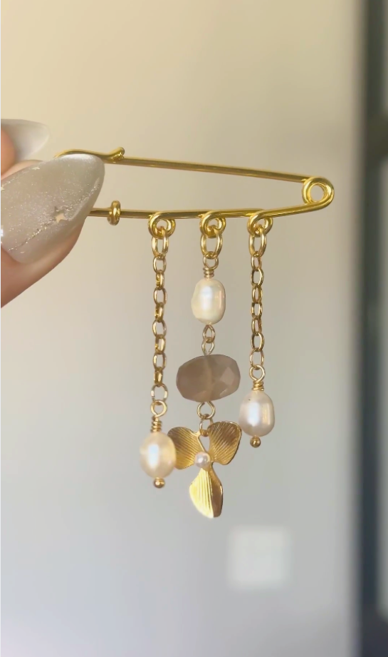 Auréad Designs Ilvara gold kilt pin brooch with peach moonstone oval, freshwater rice pearl drops, and gold three-leaf flower charm shown in motion on gold cable chain drops.